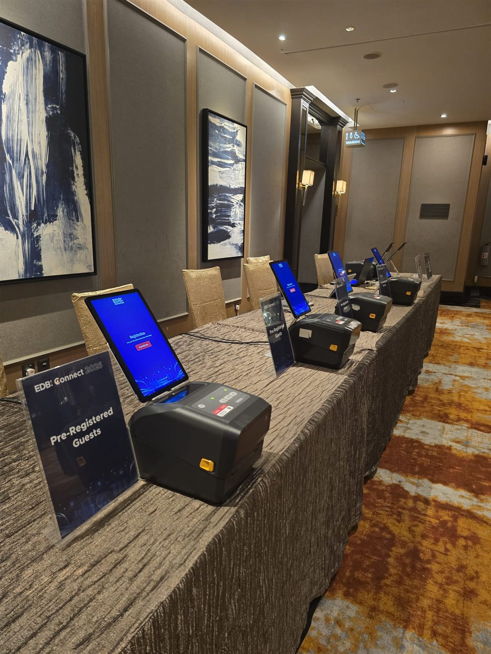 A row of tablet check-in kiosks and thermal badge printers at EDB Connect 2026